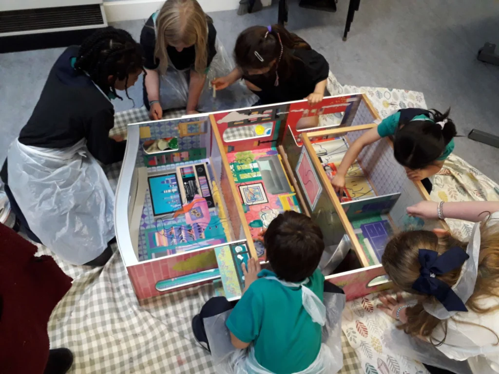 A group of children at St Joseph’s Primary School club wearing protective aprons, gathered around a large wooden dollhouse on a checkered mat, carefully decorating and arranging the interior rooms.