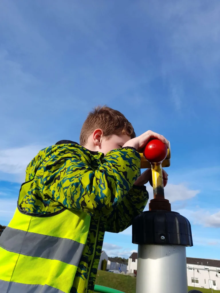 A young boy from the Dyce after school club wearing a high-visibility safety vest, pushing down on a toy pump, against a bright blue sky in Aberdeen.